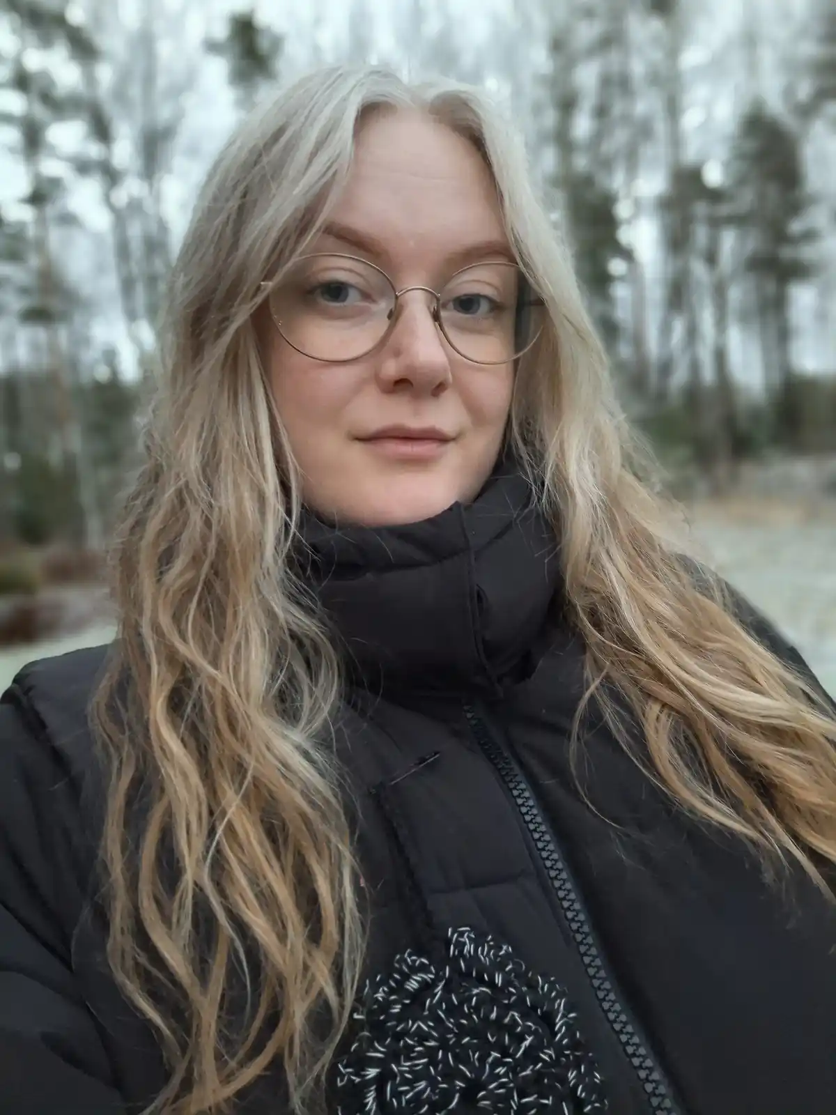 A person with long blonde hair and round glasses wears a black puffer jacket outdoors in a snowy, wooded area. The overlaid…