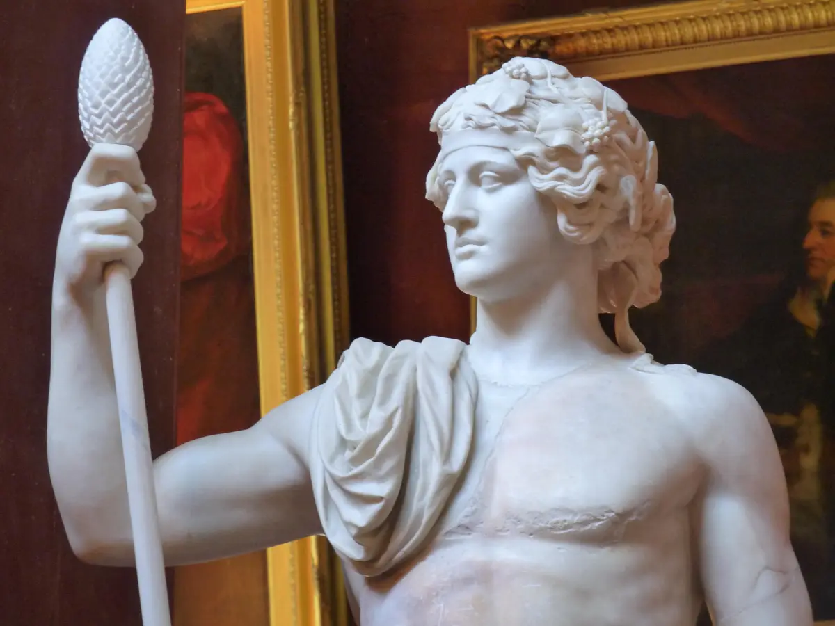 A marble statue of a classical male figure, possibly Dionysus, wearing a grapevine wreath and holding a staff topped with a…