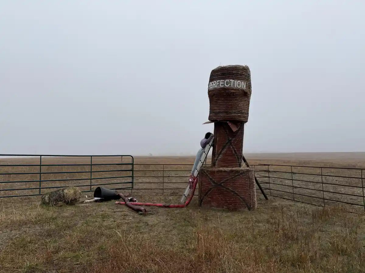 A person climbs a ladder to paint the word 'PERFECTION' on a tall stack of hay bales in a foggy field, with farm fencing and…
