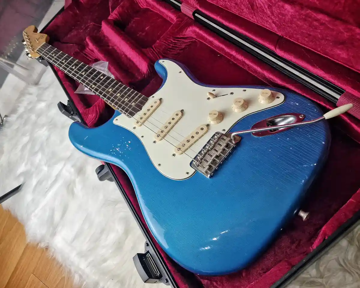 A blue electric guitar with a white pickguard and cream-colored knobs rests inside an open case lined with red velvet. The…