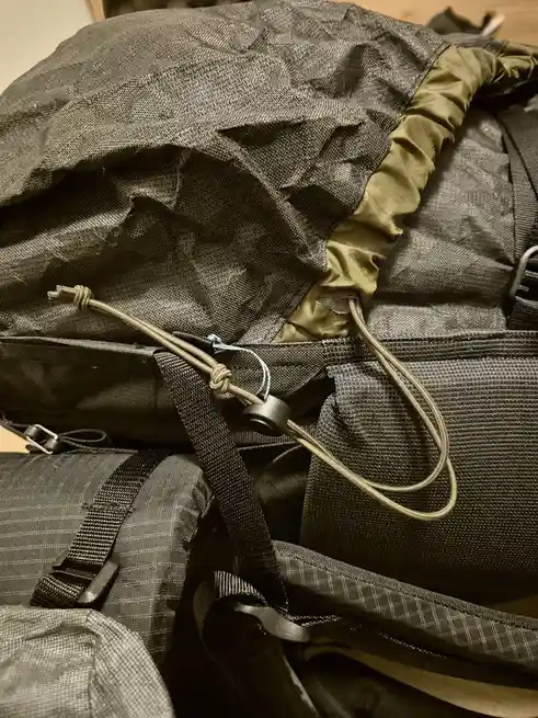 A close-up photo of a dark grey and olive green backpack with visible straps, buckles, and a drawstring closure. A small…
