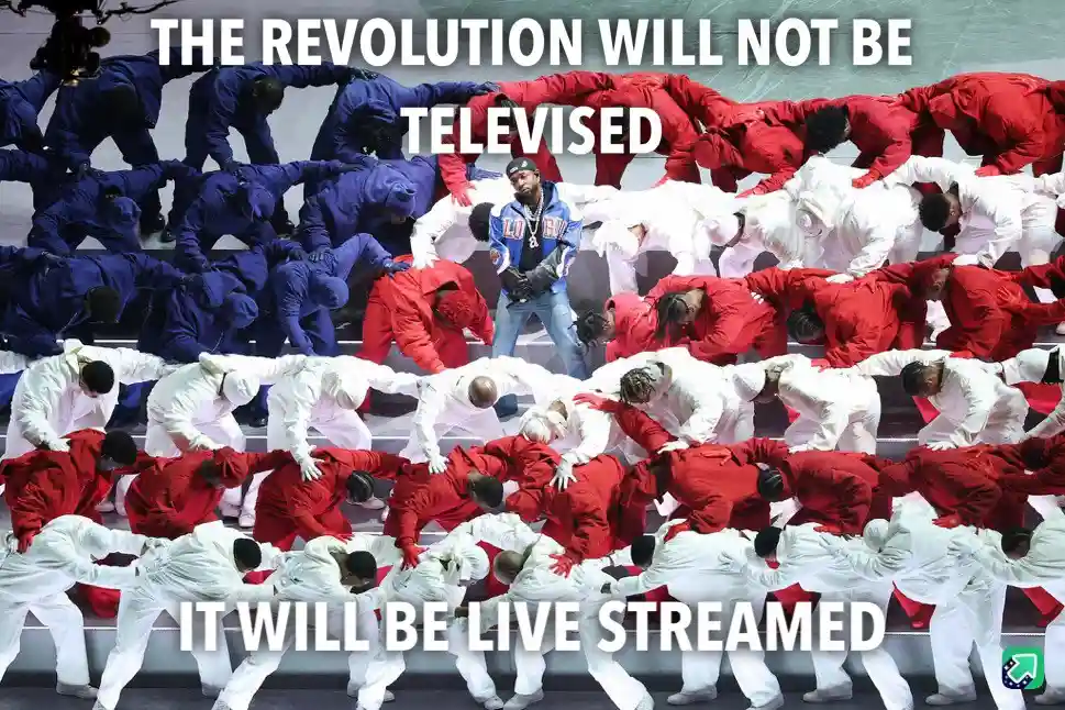 A photo of a man in a blue jacket standing center stage surrounded by rows of dancers in red, white, and blue outfits…