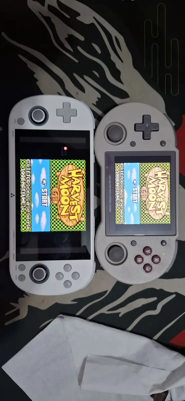 Two handheld gaming devices, a white Triniti Smart Pro and a gray handheld console, both display the title screen of the…
