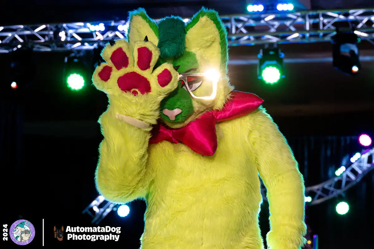 A person in a bright yellow furry costume with green accents, wearing a red bowtie and large glasses, holds up a paw with…