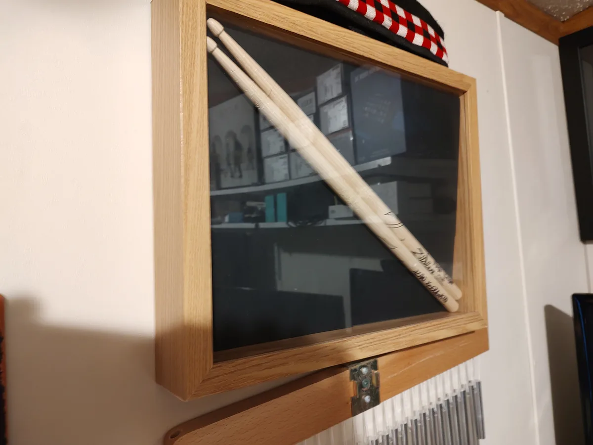 A pair of wooden drumsticks are displayed diagonally inside a light-wood framed glass case mounted on a wall, with a…