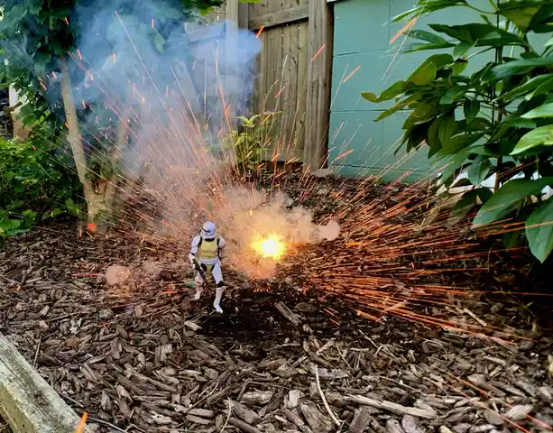 A small stormtrooper action figure stands in a garden bed covered in wood chips, facing a bright explosion that sends orange…