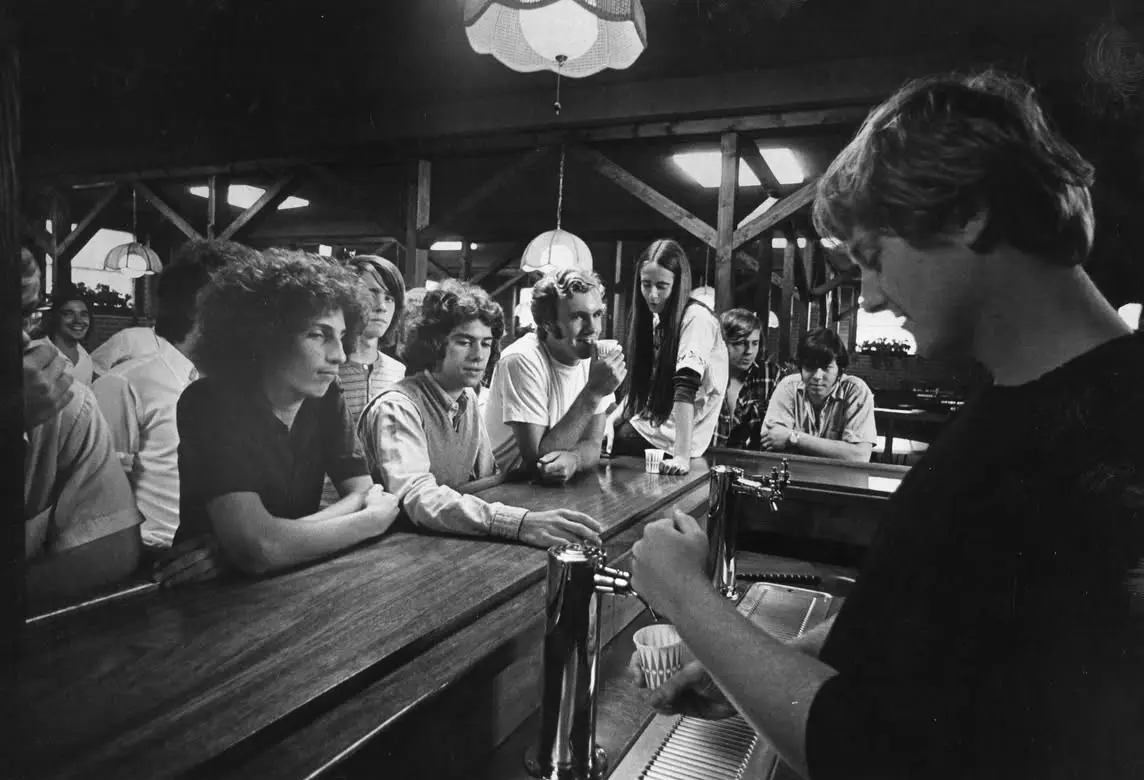 A black-and-white photo shows a young man in a dark shirt pouring a drink from a tap behind a wooden bar, while several…