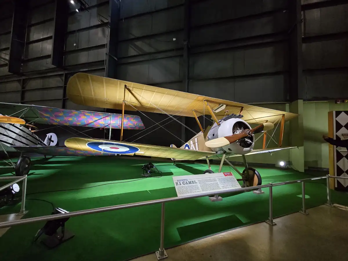 A vintage biplane, identified as a Sopwith F.1 Camel, is displayed in a museum with green flooring and metal railings. The…