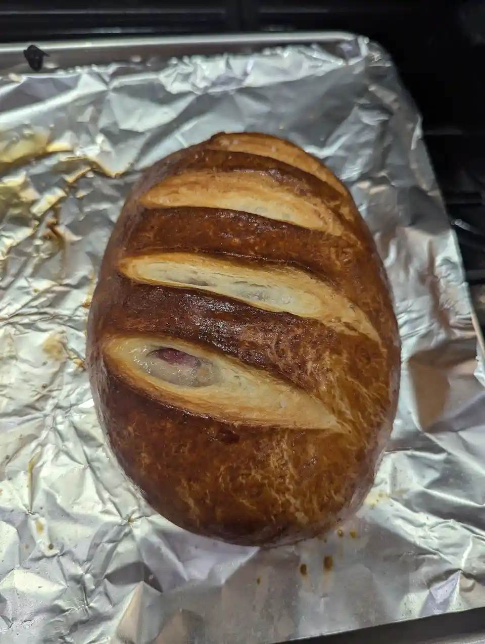 A golden-brown, oval-shaped loaf of bread sits on crumpled aluminum foil on a baking tray. The bread has diagonal slashes…