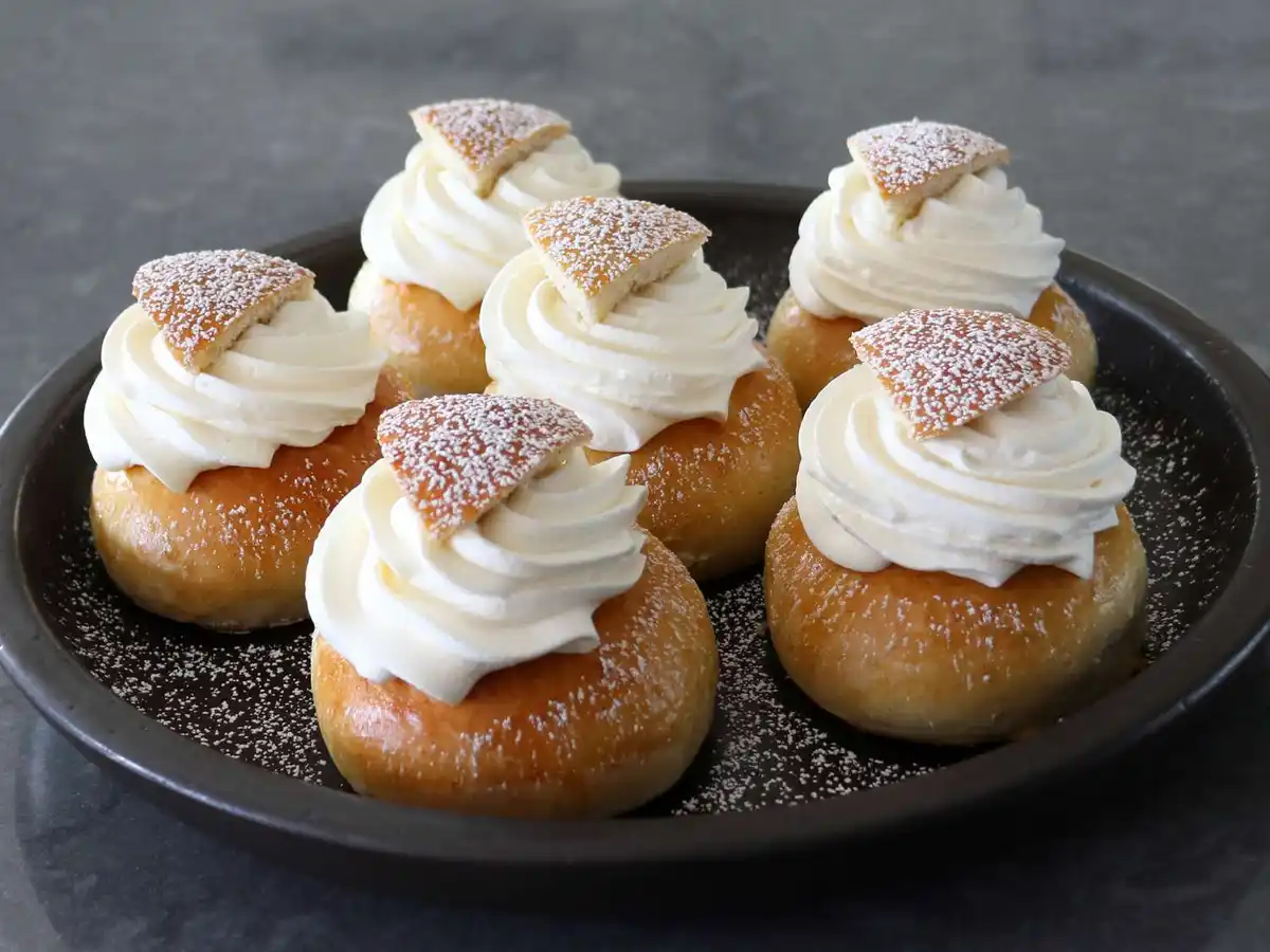 A dark plate holds six cream puffs, each topped with a swirl of white cream and a small, powdered sugar-dusted pastry piece…