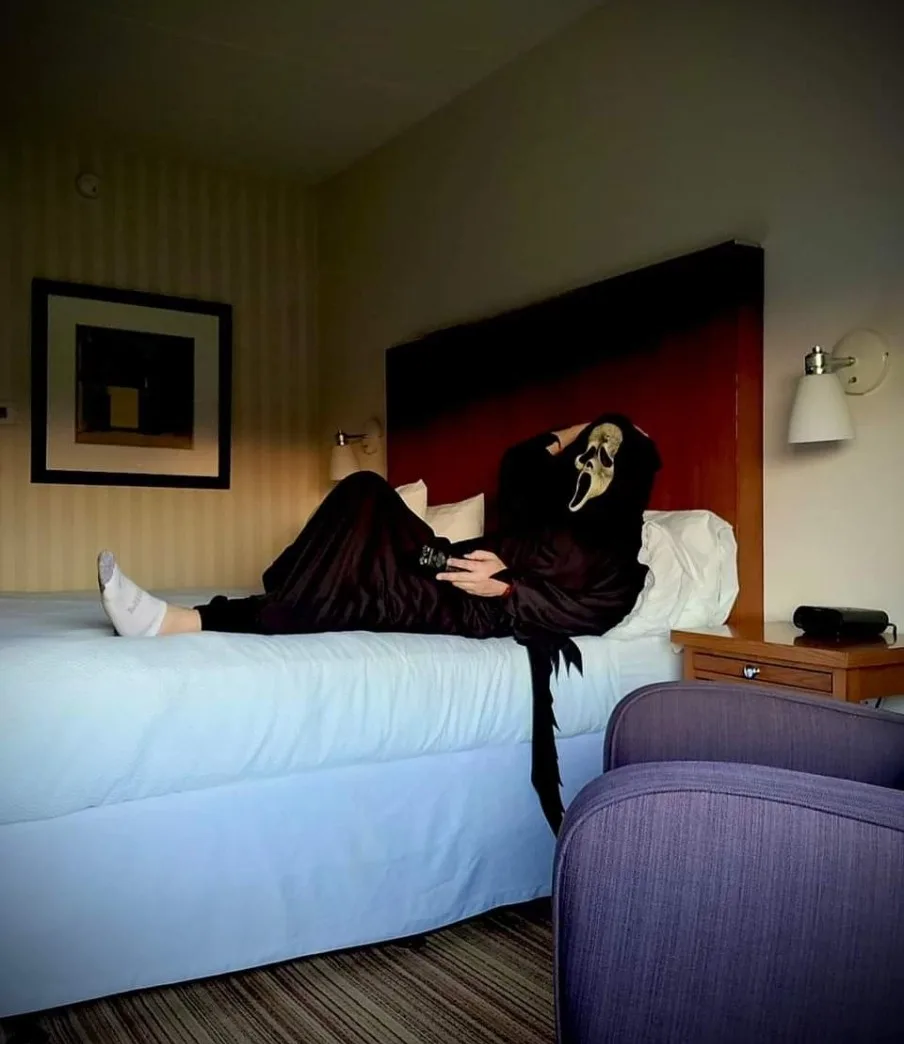 A person wearing a Ghostface mask from the 'Scream' movie franchise is lounging casually on a hotel bed, holding a phone…