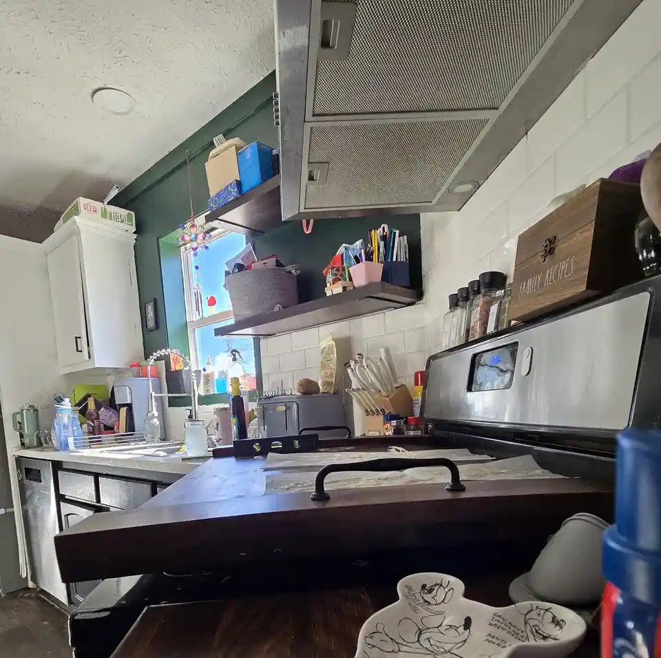 A cluttered kitchen with white cabinets, a stainless steel stove, and open shelves holding various items like spice jars, a…