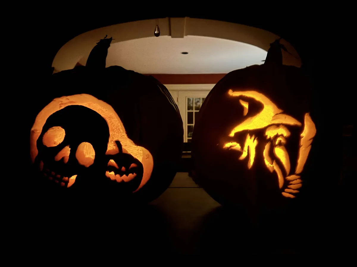 Two large, intricately carved Halloween pumpkins glow from within in a dark doorway. The left pumpkin features a skull…