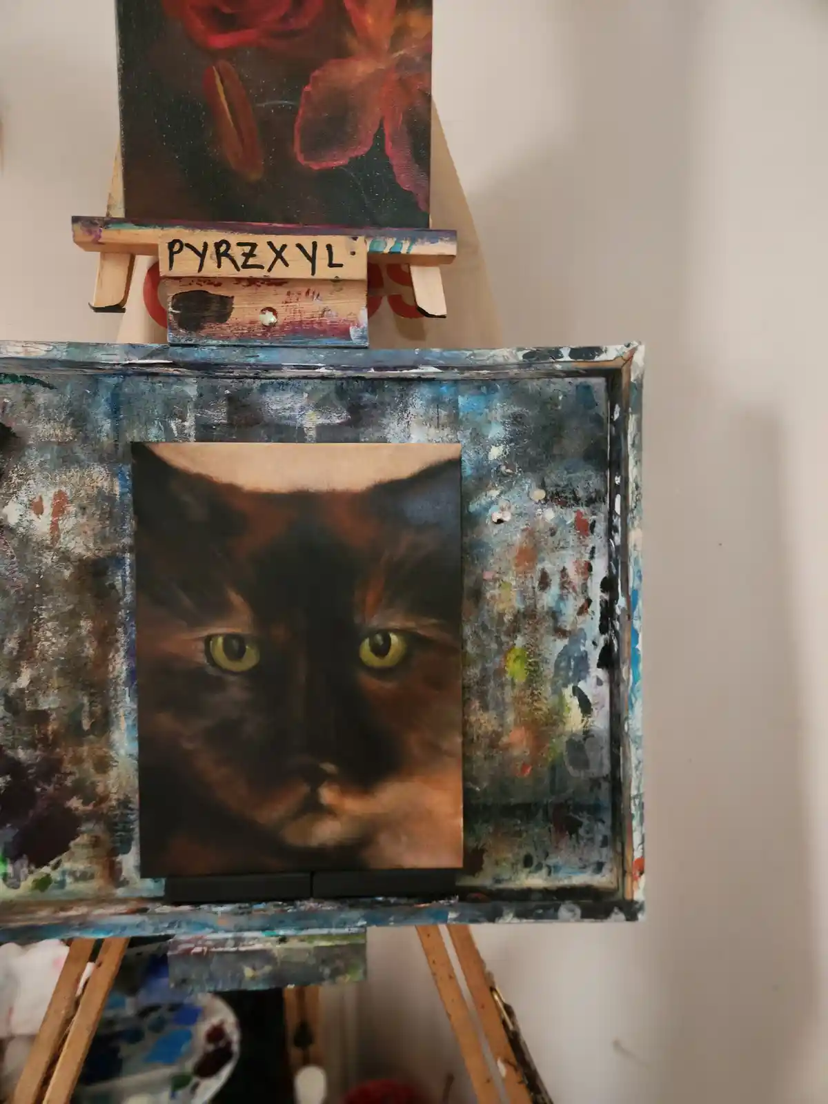 A close-up painting of a cat's face with intense yellow eyes is displayed on a paint-splattered easel. Above it, another…