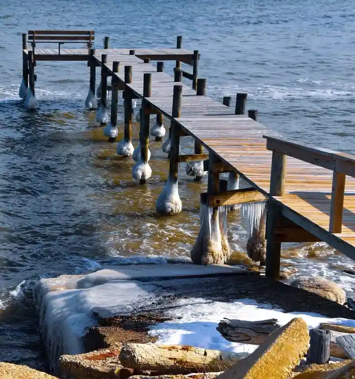 A wooden pier extends into icy water, with its support posts encased in thick, bulbous ice formations and icicles hanging…