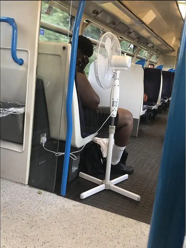 A person sits on a train seat with a white standing fan plugged into an outlet beside them, positioned to blow air directly…