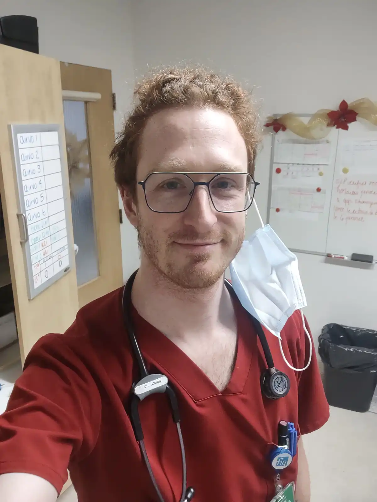 A man with red hair and glasses, wearing red scrubs, a stethoscope, and a face mask hanging from his ear, smiles at the…