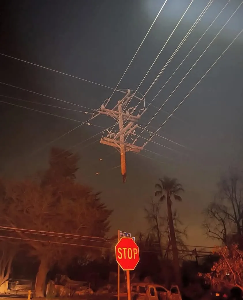 A nighttime photo shows a wooden utility pole hanging by wires from above, suspended in mid-air over a street with a visible…