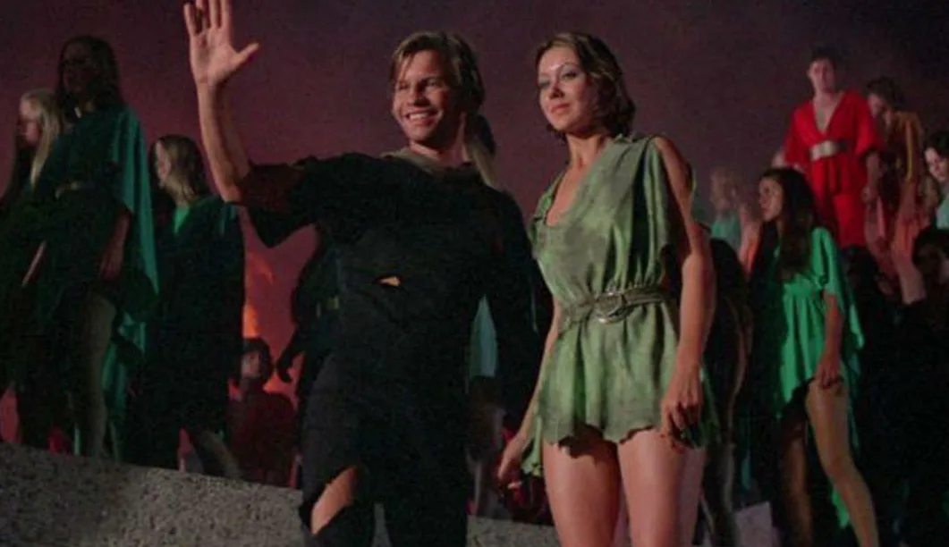 A man in a torn black outfit waves enthusiastically while standing beside a woman in a green dress, both smiling as they…
