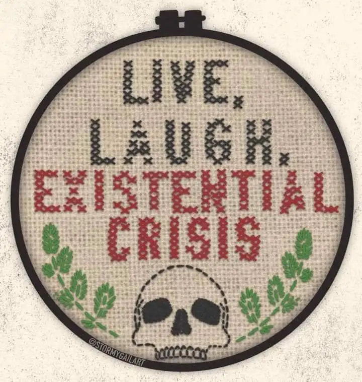 A cross-stitch pattern in a black embroidery hoop displays the text 'LIVE. LAUGH. EXISTENTIAL CRISIS' in black and red…
