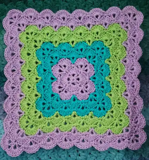 A close-up photo of a colorful, hand-crocheted square blanket with concentric rings of lavender, lime green, and teal yarn…