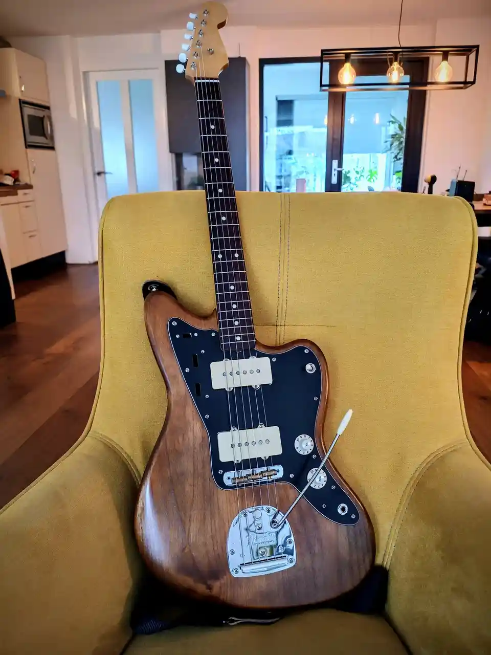 A wooden-bodied electric guitar with a black pickguard and white pickups rests upright on a mustard-yellow upholstered…