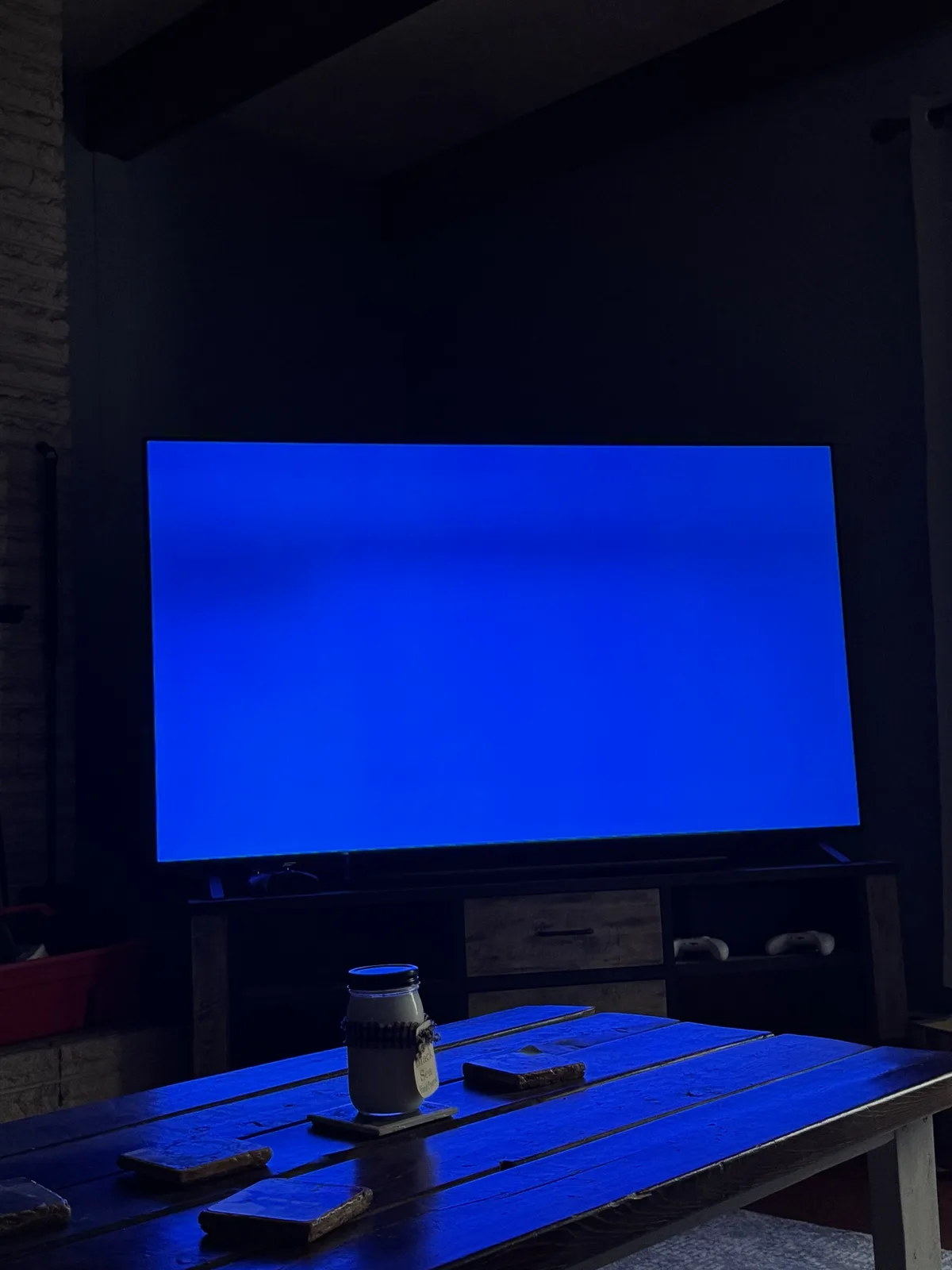 A dimly lit room features a large TV displaying a solid blue screen, casting a blue glow on a wooden table in front. On the…