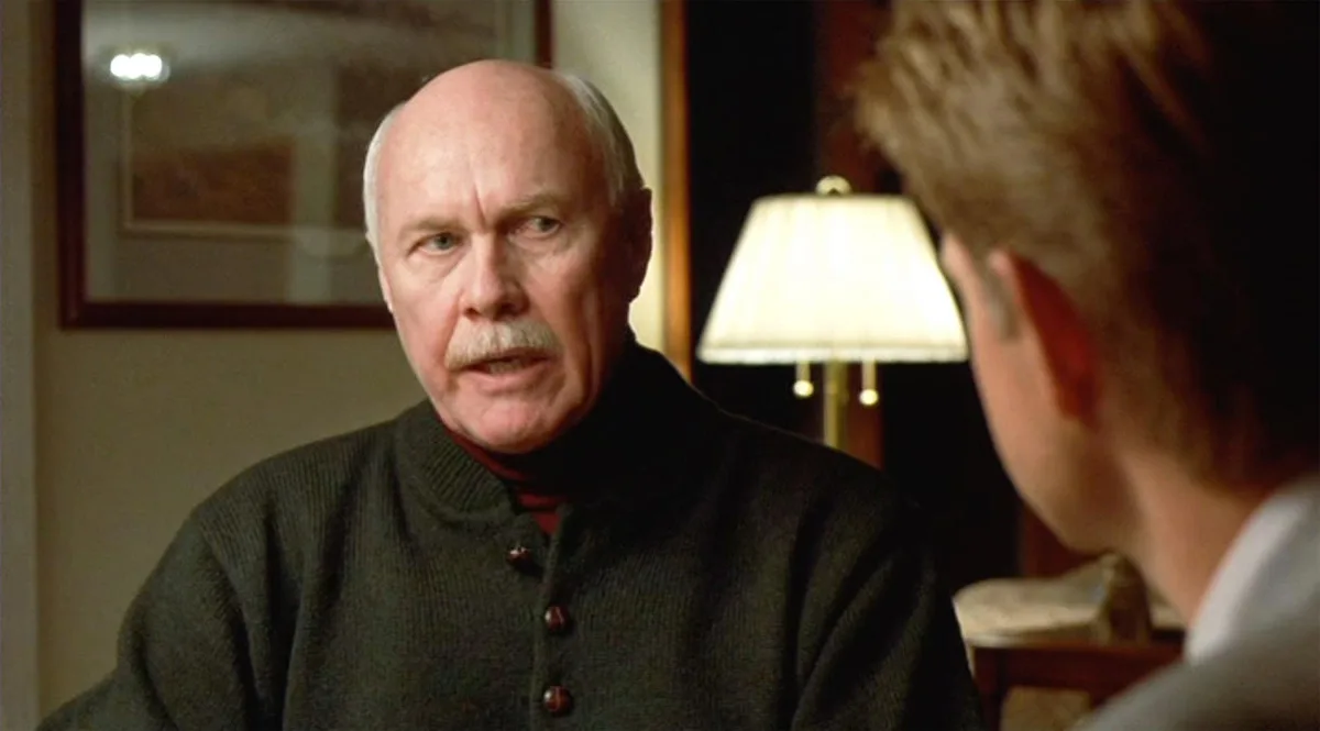 a still from a movie shows an older man with a mustache and balding head wearing a dark cardigan over a turtleneck, looking…