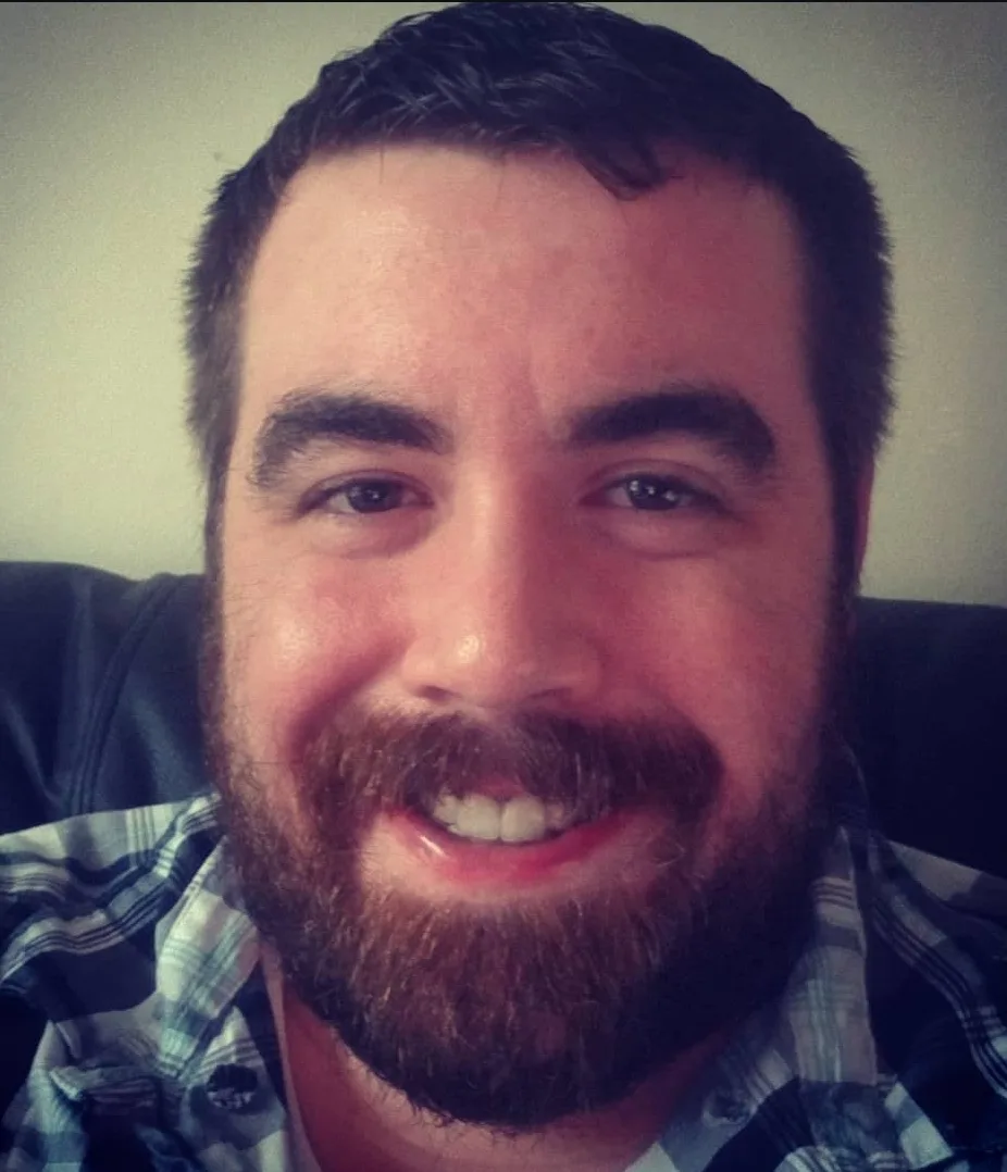 A close-up selfie of a smiling man with a full beard and short dark hair, wearing a plaid shirt. The image has a slightly…