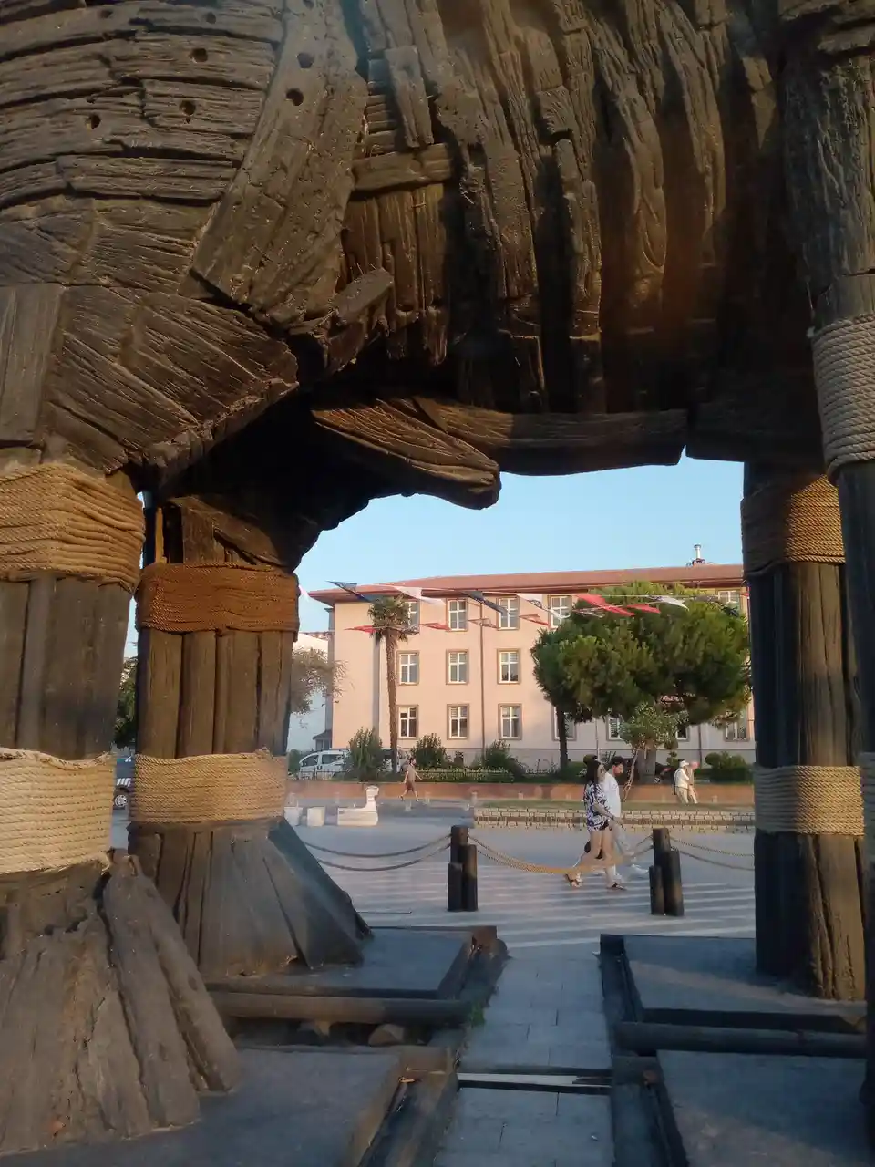 A photo shows a large, rustic wooden sculpture resembling a ship's hull or frame, with thick ropes wrapped around its posts…