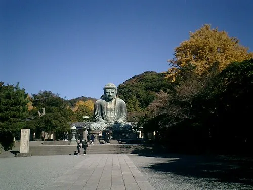 A large bronze Buddha statue sits serenely outdoors under a clear blue sky, surrounded by autumn trees with yellow and green…