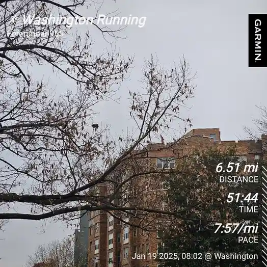 A screenshot of a Garmin running app overlay on a photo of bare tree branches and brick buildings under an overcast sky. The…