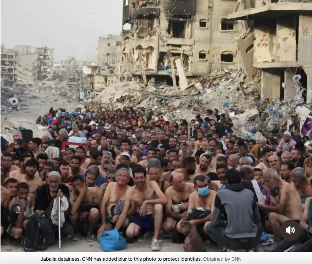 A large group of men, many shirtless and sitting or crouching on rubble, are gathered in front of heavily damaged…