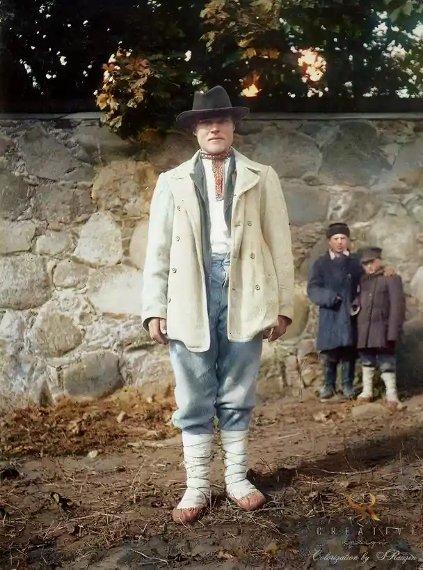 A colorized vintage photo shows a man in early 20th-century attire, including a hat, light-colored jacket, and distinctive…