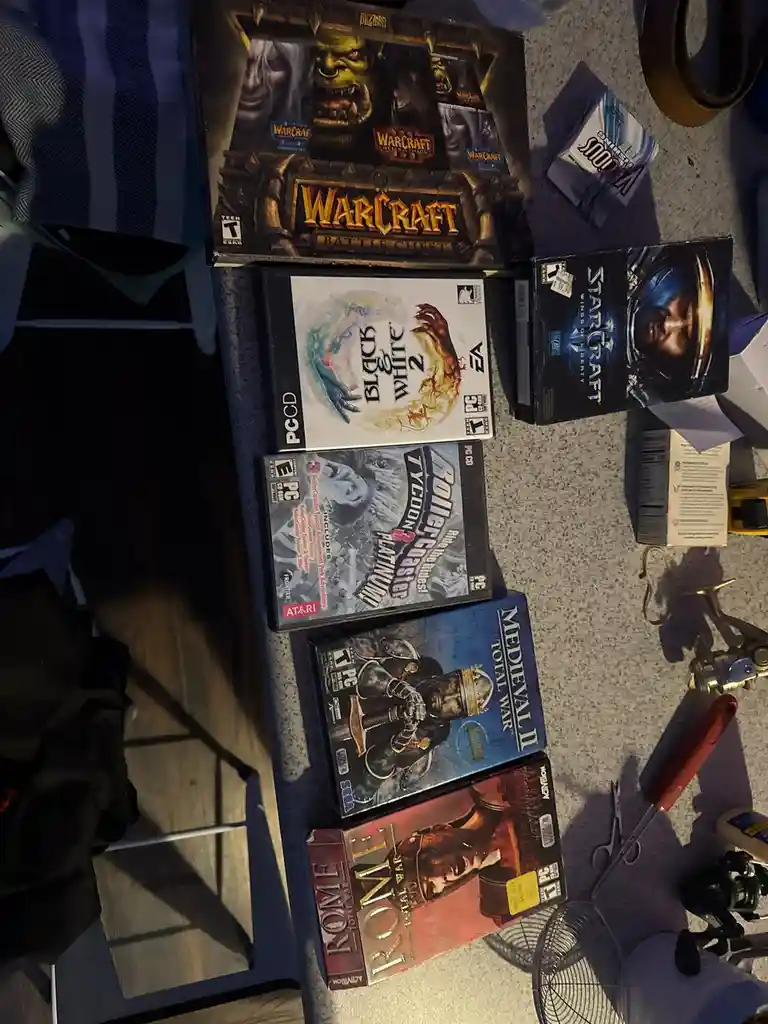 An overhead shot displays a collection of PC game boxes including Warcraft Battle Chest, Black & White 2, StarCraft: Wings…