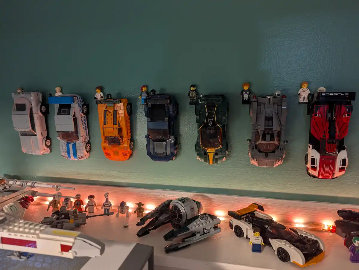 A row of seven LEGO car models are mounted upside down on a teal wall, each with a small LEGO minifigure perched on top…