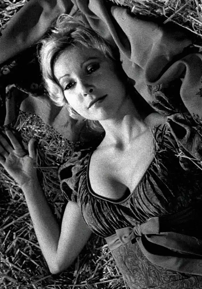 A black-and-white photograph shows a woman with blonde hair lying on her back in a field of hay, wearing a patterned dress…