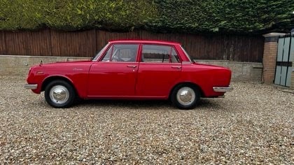 A red vintage four-door sedan is parked on a gravel driveway with a wooden fence and green hedges in the background. The car…