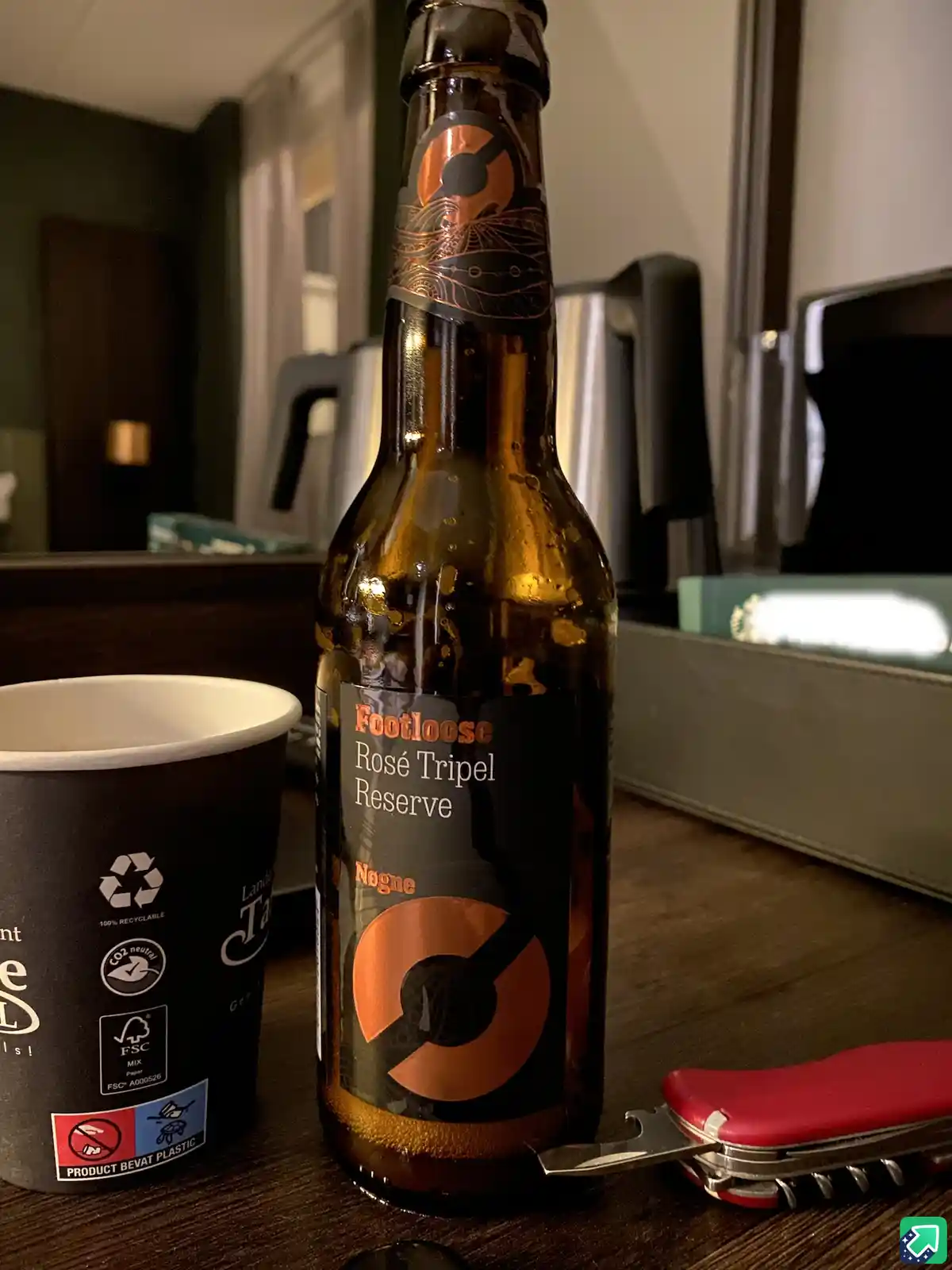 A chilled brown beer bottle labeled 'Footloose Rosé Tripel Reserve' sits on a wooden surface next to a black paper cup and a…