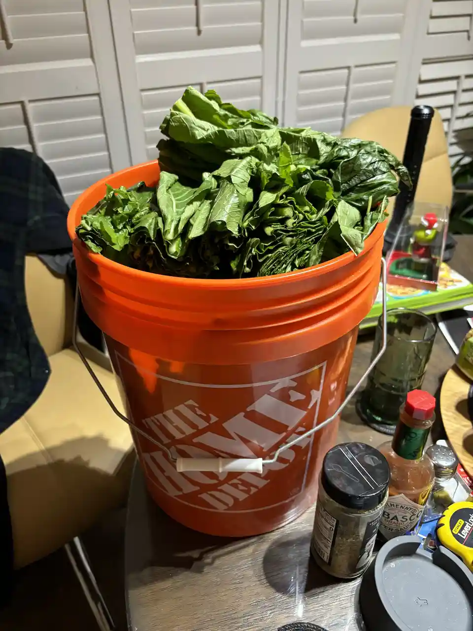 An orange Home Depot bucket overflowing with fresh green spinach leaves sits on a wooden table, surrounded by kitchen items…
