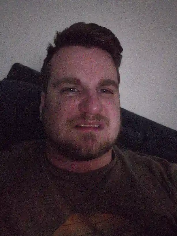 A low-resolution, grainy selfie of a man with short dark hair and a goatee, smiling slightly while seated on a dark couch…