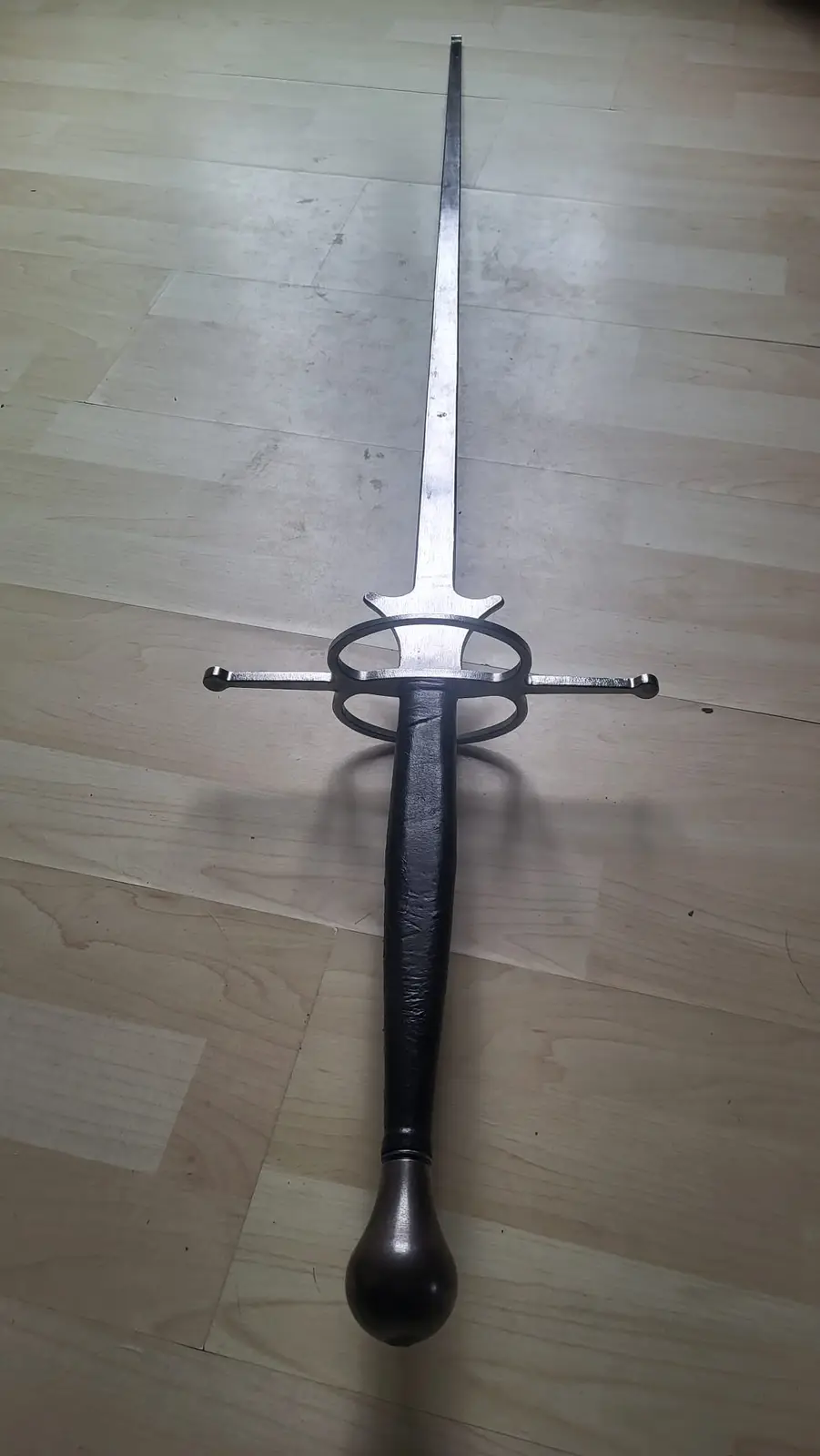 A long, silver-colored rapier-style sword with a black leather-wrapped grip and a rounded pommel lies horizontally on a…