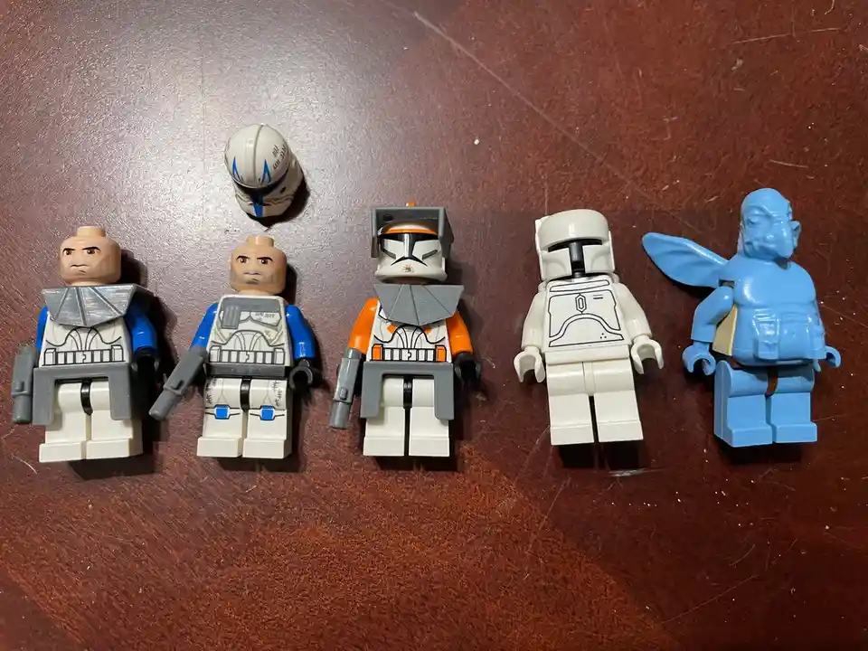 Five LEGO Star Wars minifigures are lined up on a wooden surface, four dressed as clone troopers with varying armor colors…