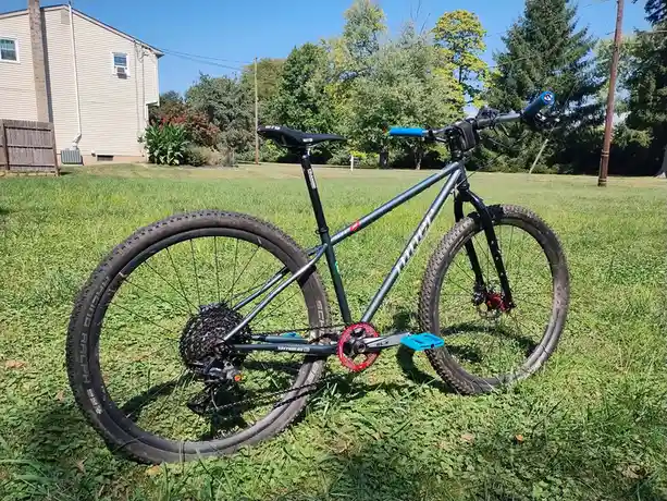 A mountain bike with a dark green frame, blue handlebars, and red crankset is parked upright in a grassy yard. A beige house…