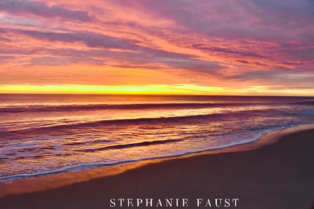 A serene beach sunset photo with vibrant orange, pink, and purple clouds over the ocean, gentle waves lapping at the shore…