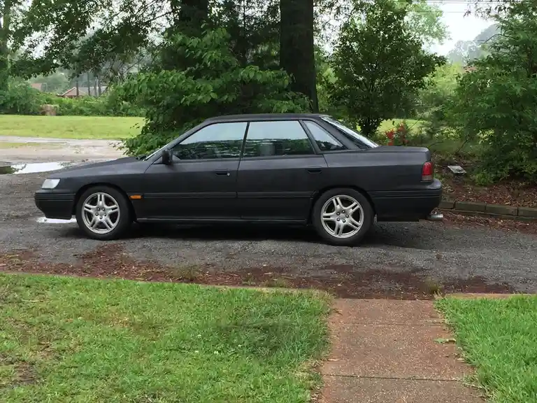 A black sedan with a matte finish is parked on a gravel driveway surrounded by green grass and trees. The car has silver…