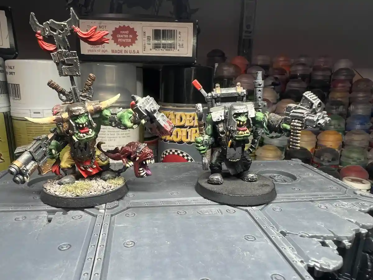 Two heavily armed, green-skinned fantasy miniatures stand on a metal surface, surrounded by paint cans and hobby supplies…