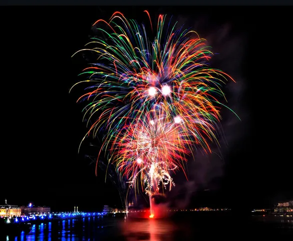 A vibrant, multi-colored fireworks display explodes over a body of water at night, with city lights and reflections visible…