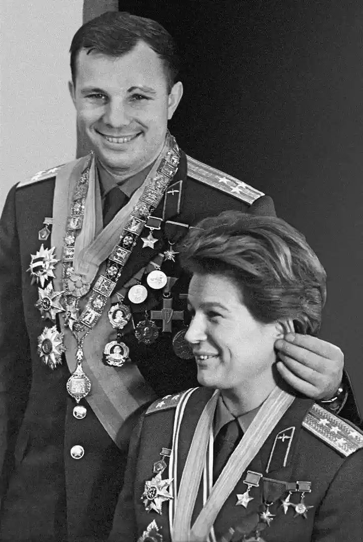 A black-and-white photo shows a man in a decorated military uniform smiling broadly while a woman in a similar uniform, also…