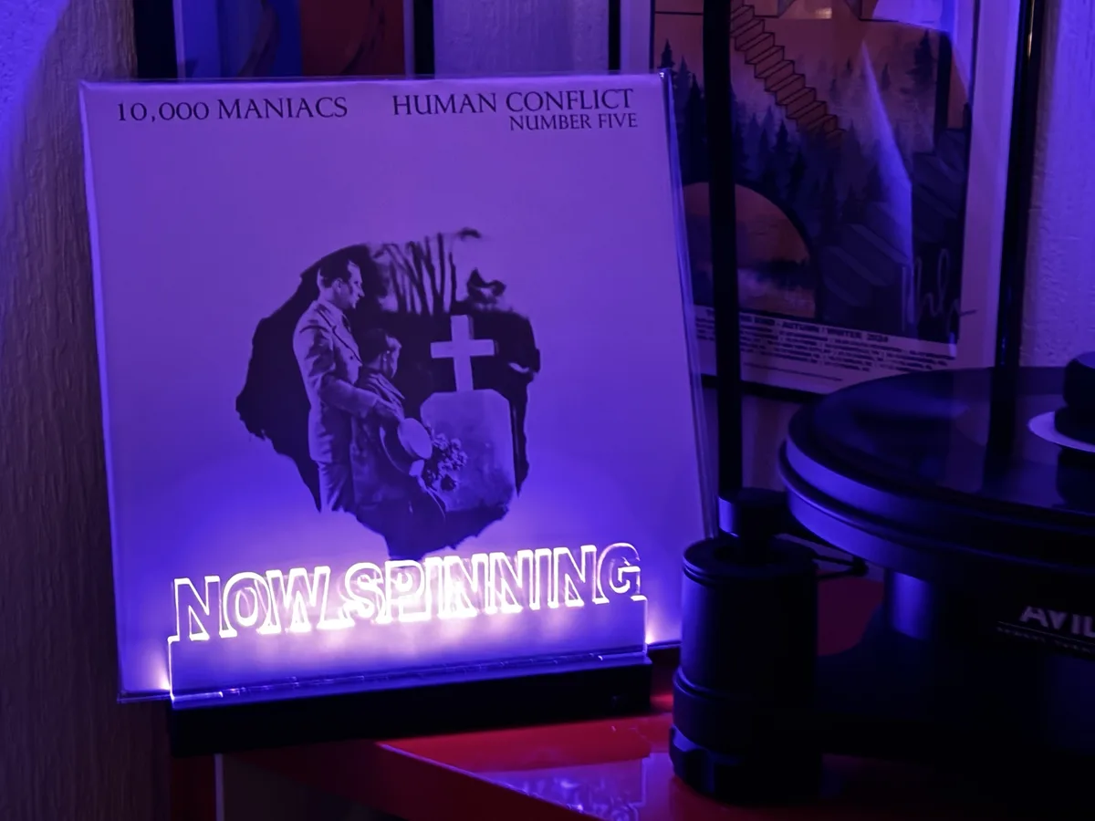 A glowing 'NOW SPINNING' sign is placed in front of a vinyl record sleeve for '10,000 Maniacs - Human Conflict Number Five'…